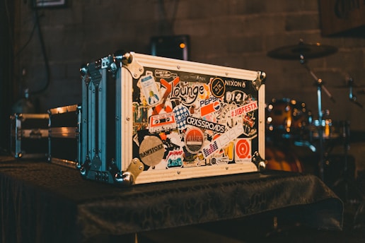 A large metal flight case covered in various colorful stickers sits on a table. The background reveals a drum set partially visible in a dimly lit room with a rustic wall, creating an ambiance often found in music venues.