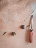 A flat surface with a corrugated texture features several metal screws and knobs scattered around. A screwdriver with a red and black handle lies on the right side of the scene.
