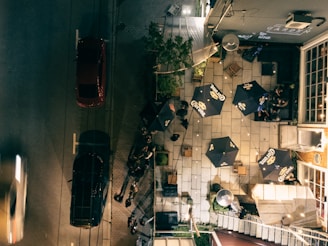 An aerial view of an outdoor patio area with several tables shaded by large promotional umbrellas. Each table appears to be occupied by people relaxing and conversing. The setting is adjacent to a street where cars are parked alongside. A combination of natural greenery and urban elements such as bricks and tiles decorates the space. The lighting suggests an evening setting, casting shadows and illuminating the seating area.