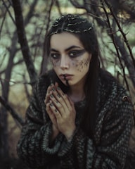 A woman with elaborate makeup and nail art is portrayed amidst a dense, barren forest setting. Her face is painted with dark shades and textures, and her nails feature similarly styled art. She wears a textured, dark garment and her hair is adorned with twigs or small branches, enhancing the natural, earthy atmosphere.