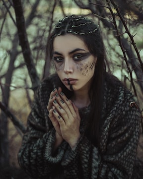 A woman with elaborate makeup and nail art is portrayed amidst a dense, barren forest setting. Her face is painted with dark shades and textures, and her nails feature similarly styled art. She wears a textured, dark garment and her hair is adorned with twigs or small branches, enhancing the natural, earthy atmosphere.