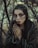A woman with elaborate makeup and nail art is portrayed amidst a dense, barren forest setting. Her face is painted with dark shades and textures, and her nails feature similarly styled art. She wears a textured, dark garment and her hair is adorned with twigs or small branches, enhancing the natural, earthy atmosphere.