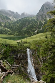 A serene landscape featuring a cascading waterfall flowing over rugged rocks, surrounded by lush greenery. In the background, mist-covered mountains rise steeply, and a tranquil lake lies nestled at the base of the hills. The scene is rich with natural beauty and untouched wilderness.