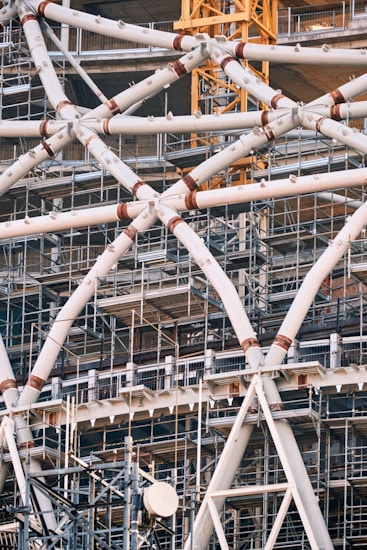 Detailed view of a complex steel structure with interconnected metal beams and support scaffolding. Numerous pipes and girders crisscross in a geometric pattern, accompanied by visible construction elements such as scaffolding and a yellow crane in the background. The scene captures an intricate industrial design, showcasing the framework of a developing building or structure.