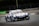 A racing car is moving at high speed on a track. The car is adorned with multiple sponsor logos, featuring a sleek design and vibrant colors. The surrounding environment includes a race track with barriers and advertisements in the background.