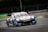 A racing car is moving at high speed on a track. The car is adorned with multiple sponsor logos, featuring a sleek design and vibrant colors. The surrounding environment includes a race track with barriers and advertisements in the background.