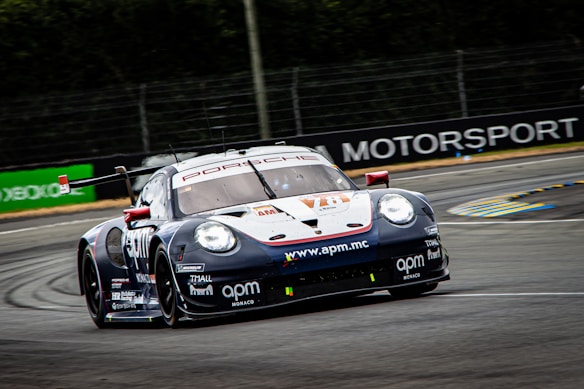 A racing car is moving at high speed on a track. The car is adorned with multiple sponsor logos, featuring a sleek design and vibrant colors. The surrounding environment includes a race track with barriers and advertisements in the background.
