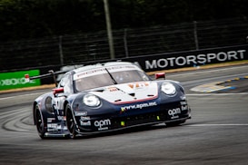 A racing car is moving at high speed on a track. The car is adorned with multiple sponsor logos, featuring a sleek design and vibrant colors. The surrounding environment includes a race track with barriers and advertisements in the background.