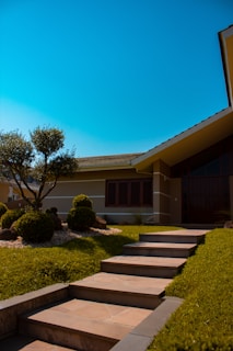 A modern house with a sloped roof and large windows, surrounded by a neatly landscaped garden featuring trimmed bushes and a small tree. The pathway to the entrance consists of evenly spaced stone steps bordered by grass, under a clear blue sky that enhances the serene ambiance.