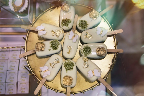Nine decorative ice cream bars arranged in a circular pattern on a golden platter. Each ice cream bar is coated in a pale green or white glaze and adorned with gold foil and decorative pieces such as edible flowers and green beads.