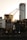 An industrial crane stands prominently against a city skyline at sunset. The crane's structure is silhouetted by the warm glow of the evening sun, highlighting its intricate metalwork. Surrounding the crane are modern skyscrapers with reflective glass facades and an older brick building, creating a contrast between industrial and urban elements.