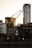 An industrial crane stands prominently against a city skyline at sunset. The crane's structure is silhouetted by the warm glow of the evening sun, highlighting its intricate metalwork. Surrounding the crane are modern skyscrapers with reflective glass facades and an older brick building, creating a contrast between industrial and urban elements.