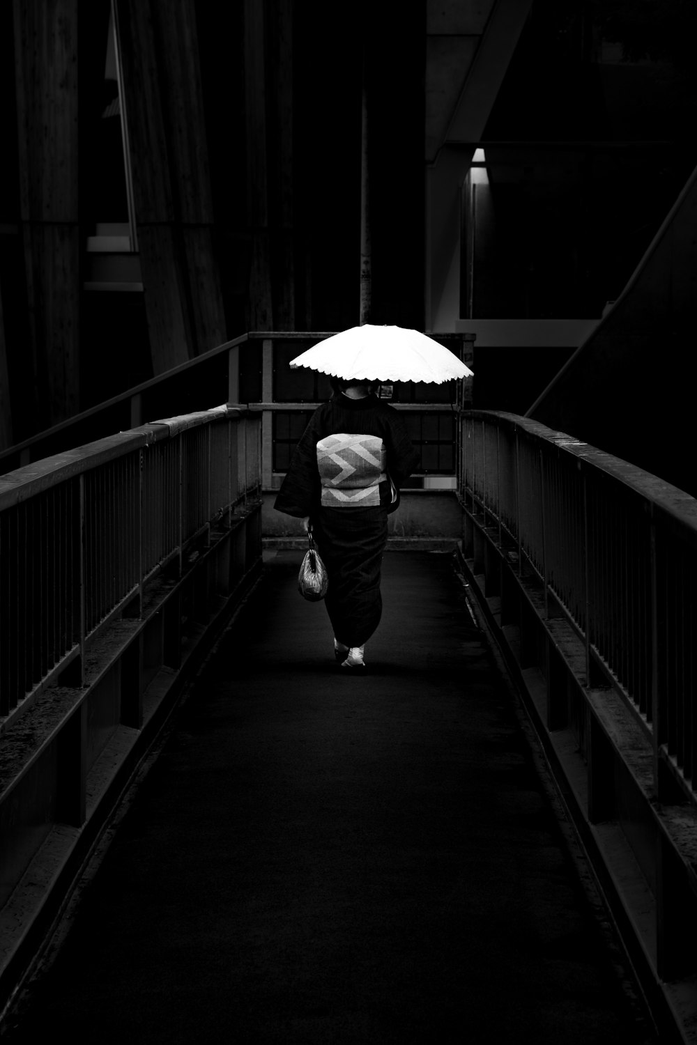 grayscale photo of man in jacket holding umbrella walking on bridge