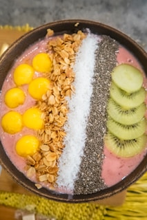 A vibrant smoothie bowl features an array of toppings arranged in neat rows. On the left, bright yellow balls of mango rest against a pink smoothie base. Next to the mango, a line of golden brown granola provides a crunchy texture. The center is decorated with a strip of white shredded coconut, followed by a layer of tiny black chia seeds. On the right, sliced green kiwis complete the colorful display.