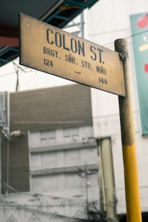 A weathered street sign for Colon St. with directions and numbers, mounted on a pole with urban buildings and electrical wires visible in the background.
