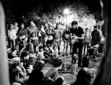 A street performer engages a gathered crowd, including both adults and children. The audience is seated on the ground and standing, fixated on the performer who appears to be controlling a puppet. The scene is set against a backdrop of trees illuminated by overhead streetlights, creating a lively night atmosphere.