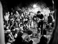 A street performer engages a gathered crowd, including both adults and children. The audience is seated on the ground and standing, fixated on the performer who appears to be controlling a puppet. The scene is set against a backdrop of trees illuminated by overhead streetlights, creating a lively night atmosphere.