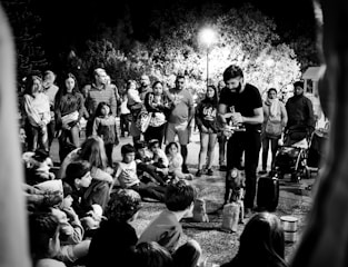 A street performer engages a gathered crowd, including both adults and children. The audience is seated on the ground and standing, fixated on the performer who appears to be controlling a puppet. The scene is set against a backdrop of trees illuminated by overhead streetlights, creating a lively night atmosphere.