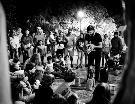 A street performer engages a gathered crowd, including both adults and children. The audience is seated on the ground and standing, fixated on the performer who appears to be controlling a puppet. The scene is set against a backdrop of trees illuminated by overhead streetlights, creating a lively night atmosphere.