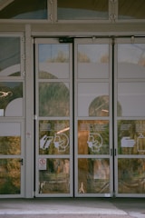 A set of glass double doors with white window decals showing instructions for hand sanitization and mask-wearing. The exterior environment, including trees and a walkway, is reflected on the glass. Hinges and door handles are visible, as well as some text printed on the doors.