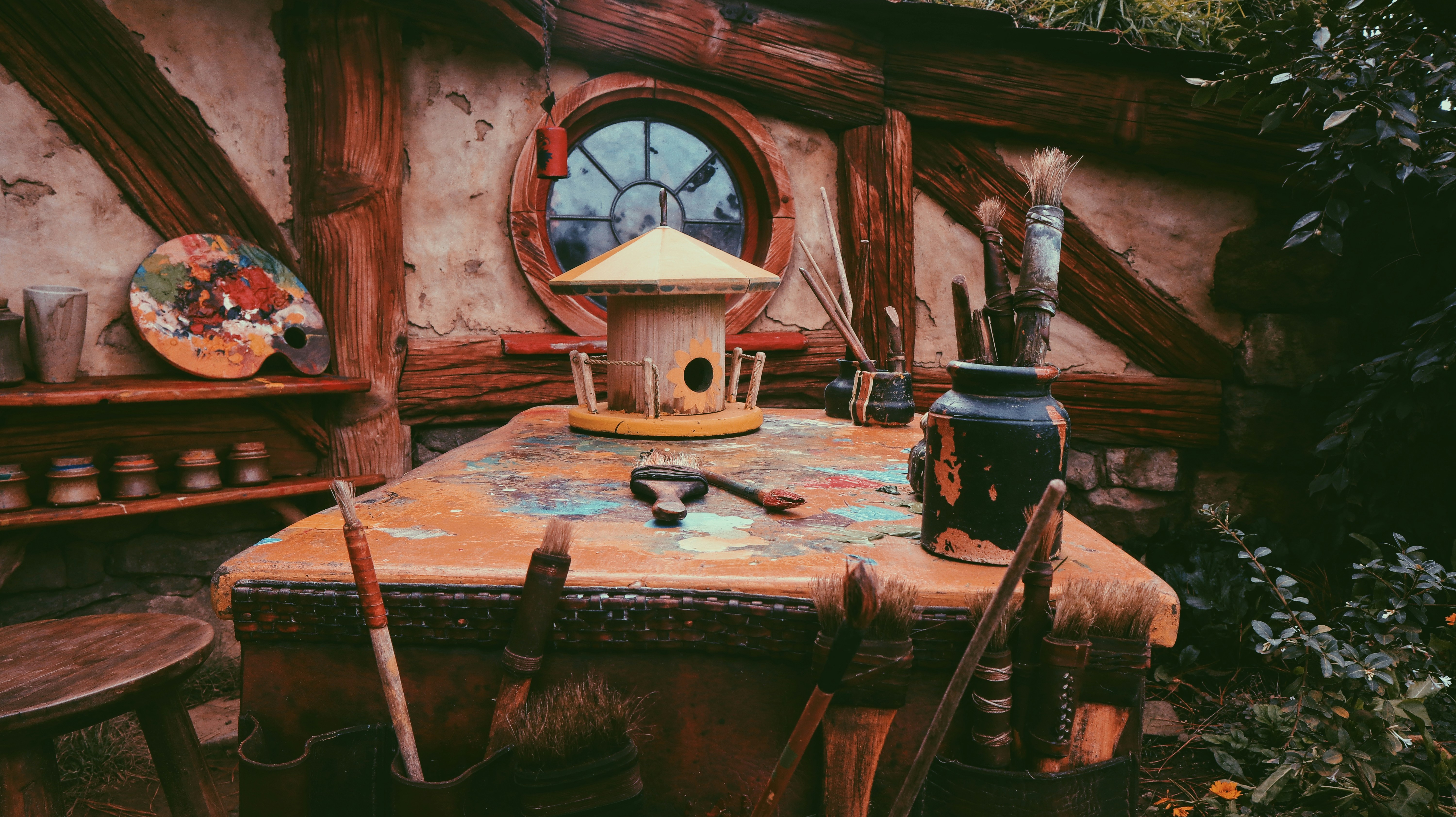 A rustic wooden table with paint and art supplies is set in a quaint outdoor setting. The table is cluttered with paintbrushes, a weathered paint palette, and a birdhouse centerpiece. The surroundings include a wooden wall with a circular window, and several clay pots are arranged on shelves.
