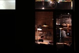 A cozy interior of a coffee shop with warm lighting, brick walls, and a person sitting at a table using a laptop. Hanging lights illuminate the room, and shelves filled with bottles and books can be seen. Another person appears to be working at the counter.