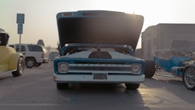 A vintage Chevrolet truck is parked with its hood open, surrounded by other classic cars. The scene is set in a parking lot during the day, with a clear sky and sunlight casting long shadows on the ground. The truck is predominantly blue with chrome details.