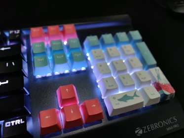 A mechanical keyboard displaying colorful keycaps with illuminated backlighting. The keys are a mix of pastel pink, blue, and white, with some featuring designs like a shark. The keyboard branding 'Zebronics Max Pro' is visible in the corner.