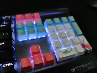 A mechanical keyboard displaying colorful keycaps with illuminated backlighting. The keys are a mix of pastel pink, blue, and white, with some featuring designs like a shark. The keyboard branding 'Zebronics Max Pro' is visible in the corner.
