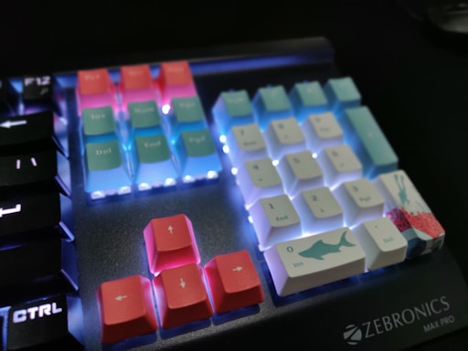 A mechanical keyboard displaying colorful keycaps with illuminated backlighting. The keys are a mix of pastel pink, blue, and white, with some featuring designs like a shark. The keyboard branding 'Zebronics Max Pro' is visible in the corner.