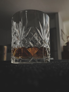 A crystal glass with intricate patterns contains a dark amber liquid, likely a type of whiskey or scotch. The setting is dimly lit, casting shadows and reflections on the glass’s surface, creating a moody atmosphere.