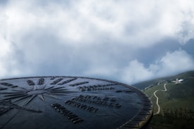 A metal compass disc with engraved text and directional arrows is prominently displayed against a backdrop of cloudy skies and a distant landscape featuring a winding path leading to a small white building.