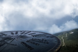 A metal compass disc with engraved text and directional arrows is prominently displayed against a backdrop of cloudy skies and a distant landscape featuring a winding path leading to a small white building.