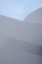 Smooth, curved architectural forms composed of subtle, overlapping layers create a sense of harmony against a clear blue sky. The minimalist design emphasizes clean lines and geometric precision, with a focus on texture and light.