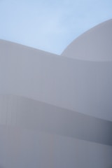 Smooth, curved architectural forms composed of subtle, overlapping layers create a sense of harmony against a clear blue sky. The minimalist design emphasizes clean lines and geometric precision, with a focus on texture and light.