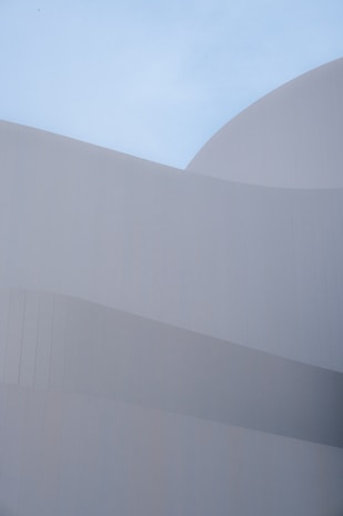 Smooth, curved architectural forms composed of subtle, overlapping layers create a sense of harmony against a clear blue sky. The minimalist design emphasizes clean lines and geometric precision, with a focus on texture and light.