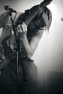 A musician is intensely playing an electric guitar on stage, leaning over with their hair hanging down and wearing sunglasses. The lighting creates a dramatic effect, casting shadows over the musician, who is wearing a casual t-shirt and shorts. A microphone stand is visible in the foreground.