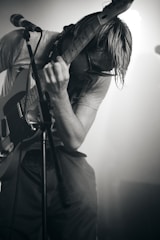 A musician is intensely playing an electric guitar on stage, leaning over with their hair hanging down and wearing sunglasses. The lighting creates a dramatic effect, casting shadows over the musician, who is wearing a casual t-shirt and shorts. A microphone stand is visible in the foreground.