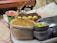 A variety of food items are arranged in a rustic setting. There are sacks of grains, trays of chopped tomatoes, green chilies, lemons, and a pile of roasted chickpeas. The items are resting on a makeshift table covered with a light cloth, with metal bowls and a woven basket visible.