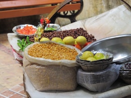 A variety of food items are arranged in a rustic setting. There are sacks of grains, trays of chopped tomatoes, green chilies, lemons, and a pile of roasted chickpeas. The items are resting on a makeshift table covered with a light cloth, with metal bowls and a woven basket visible.