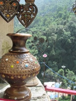 A decorative, intricately carved wooden vase with colorful decorative elements is placed on a ledge. Above it, two heart-shaped metal ornaments with floral patterns hang. In the background, dense green foliage covers the hilly landscape, creating a lush, natural setting.