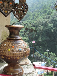 A decorative, intricately carved wooden vase with colorful decorative elements is placed on a ledge. Above it, two heart-shaped metal ornaments with floral patterns hang. In the background, dense green foliage covers the hilly landscape, creating a lush, natural setting.