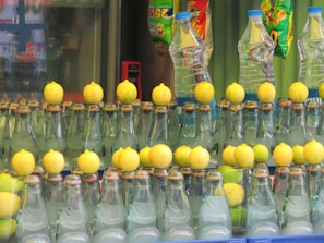 A row of glass bottles filled with a clear liquid, each topped with a lemon. In the background, there are some plastic water bottles and colorful snack packages hanging.