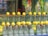 A row of glass bottles filled with a clear liquid, each topped with a lemon. In the background, there are some plastic water bottles and colorful snack packages hanging.