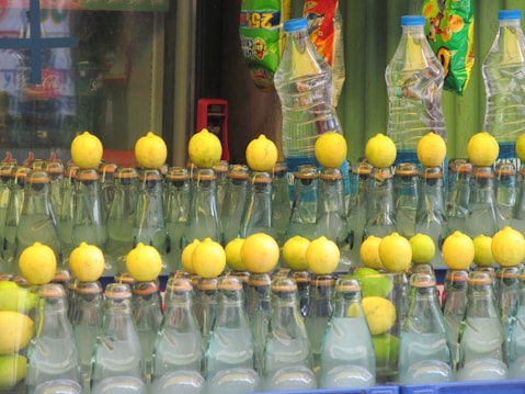 A row of glass bottles filled with a clear liquid, each topped with a lemon. In the background, there are some plastic water bottles and colorful snack packages hanging.