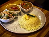 A plate featuring a serving of white rice garnished with a lemon wedge and a green leaf, accompanied by three bowls containing various dishes including a curry with red chili and another sauce with vegetables, and a small salad with shredded cabbage and carrots.