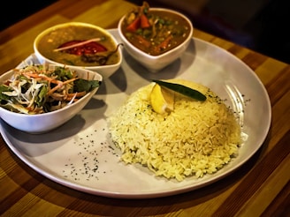 A plate featuring a serving of white rice garnished with a lemon wedge and a green leaf, accompanied by three bowls containing various dishes including a curry with red chili and another sauce with vegetables, and a small salad with shredded cabbage and carrots.