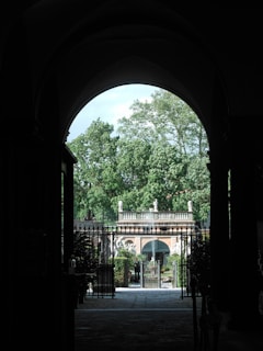 A grand archway opens to reveal an elegant garden courtyard framed by tall, lush green trees. A historic stone building is visible in the background with decorative statues adorning its roof. The courtyard is enclosed by an ornate metal gate with detailed ironwork.