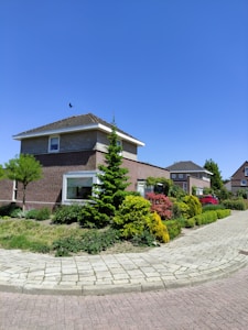 A suburban street features a neatly maintained house with a tiled roof and brick walls surrounded by a well-kept garden. The sky is clear and blue, and a bird is flying above the house. Green bushes and trees line the pathway, which is paved with bricks. Another house is visible in the background.