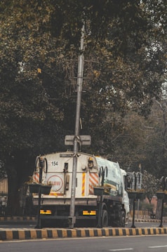 A garbage truck parked on the side of a road with large trees overhead. The road is lined with a black and yellow striped barrier. There are several potted plants attached to the truck.